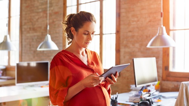 Article thumbnail: Pregnant businesswoman using tablet PC. Female professional is standing at desk. She is in brightly lit office.