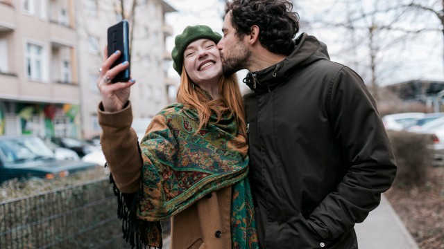 Article thumbnail: A man kissing his girlfriend on the cheek as she takes a selfie of the two of them while out for a walk in the city together.