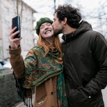 Article thumbnail: A man kissing his girlfriend on the cheek as she takes a selfie of the two of them while out for a walk in the city together.